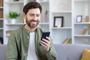 Satisfied mature man sitting on sofa in living room, using app on phone, freelancer holding smartphone, reading and typing text message, browsing social media. photo