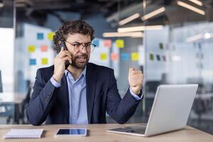 Businessman showing frustration during phone call in modern office environment. Man in suit expressing anger while working on laptop, with digital tablet and notebook on desk. photo