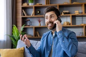 A casually dressed man in a blue shirt gestures as he speaks on a mobile phone in a modern living room setting. photo