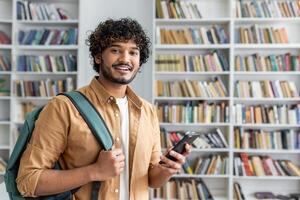 A cheerful young man with curly hair and a backpack is standing in a library. He is casually dressed, smiling,using a smartphone, possibly searching for information or communicating with friends. photo