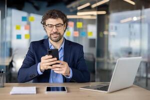 Businessman successful man with phone in hands sitting at desk inside office, mature experienced boss smiling browsing social media, typing text message, call. photo