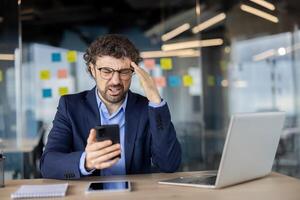 Upset and disappointed man with phone in hands at workplace, mature experienced businessman in business suit sad reading online message and checking email mail using app on smartphone. photo
