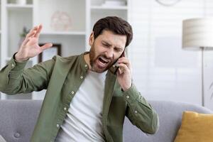 Mad unshaved man talking by cellphone and shouting angrily while raising hand in living room. Emotional guy having stressful conversation with technical support agent and enraging with bad service. photo