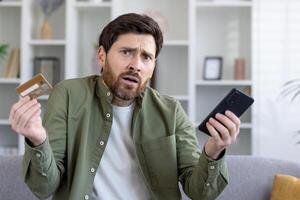 Confused adult man holding smartphone and bank card in hands while shrugging with worried expression. Uncertain customer having struggles with using ecommerce banking app for making online payments. photo