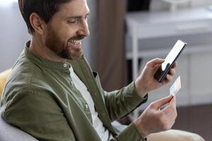 Close-up of a man sitting at home on the sofa, using an application on the phone and a bank credit and debit card, for online shopping on the Internet, smiling with satisfaction, choosing gifts. photo