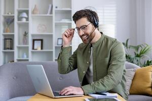Confident man working from home using a laptop and headset, smiling pleasantly. Home office setup with a cozy background. Concept of remote work and online communication. photo