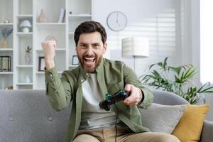 Bearded man enthusiastically playing games on a couch in a stylish, well-lit living room. He is celebrating a victory while holding a game controller. photo