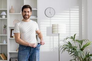 Cheerful adult male demonstrates significant weight loss by fitting into oversized jeans inside a bright modern home setting. photo