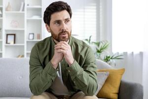 Thoughtful man sitting on a sofa in a modern living room, deep in contemplation. Bright and airy home interior with bookshelf and plants. photo