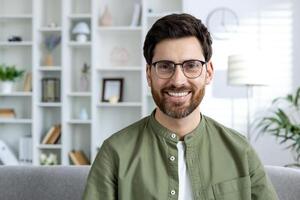 Casually dressed man with glasses smiling at home, sitting on a couch with a well-organized bookshelf behind him photo