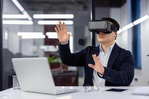A man in a business suit uses augmented reality glasses, a businessman inside an office at a workplace sits at a table with a laptop, looks at financial reports online. photo