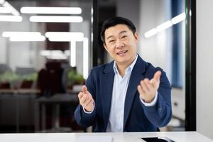 A young Asian man in a business suit is sitting in a modern office at a table in front of the camera, talking on a link, gesturing with his hands. photo