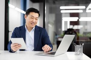 Modern businessman in formal clothes using digital tablet while working by pc on white table. Successful entrepreneur using two wireless devices for comparing articles from different web sources. photo