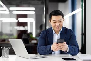 Asian man inside office with phone in hands, smiling contentedly, businessman using smartphone sitting at desk, mature boss in business suit browsing social media, typing text message call. photo