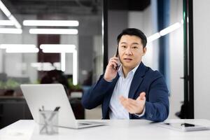 Adult entrepreneur having phone conversation while sitting bu equipped table with computer in corporate building. Calm startup leader discussing project details with perspective investor on distance. photo