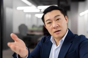 Serious asian business man looking at web cam of mobile phone and raising hand during conference. Confident male calling to distant employees in virtual chat for discussing emergency questions. photo