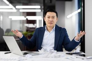 Serious office administrator raising arms aside while sitting by overloaded table with crumpled papers and pc. Busy male struggling with amount of work while coming back to office after vacation. photo