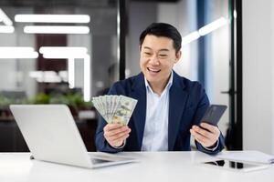 Professional businessman in a suit displaying cash and using a smartphone at a modern office work station. photo