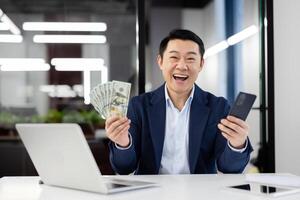 Joyful businessman in suit at office desk laughing with money in hand and a smartphone, expressing success and wealth. photo