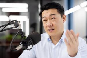 Close-up photo of an Asian young man sitting in the office at a desk in front of a microphone and having an online meeting via call, discussing, explaining, gesturing with his hand.