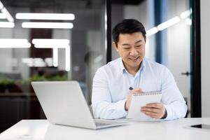 A cheerful Asian businessman is jotting down notes in a spiral notebook while working on a laptop in a contemporary office space. photo