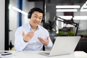 Smiling young Asian man wearing headphones sitting in office in front of desk with microphone and talking on call on laptop gesturing with hands. photo