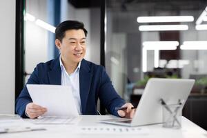 Happy person in formal clothes working by table with pc on background of modern office interior. Busy administrator accomplishing list of daily tasks and sending email with project report. photo