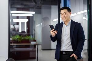 Asian businessman standing inside office with phone in hands, boss in business suit typing text message and reading social media, mature man using app on smartphone. photo