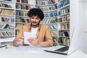 Young successful student studying sitting inside university academic library, man smiling holding tablet computer, Hispanic man using study app. photo