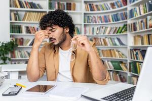 Tired muslim man taking of glasses and rubbing eyes with closed eyes while working by laptop. Stressed retired manager waiting for break while having vision struggles due to long screen time. photo