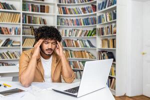Burnout latino male touching temples with arms during remote workday in front of open laptop indoors. Stressed designer having mental problems and making pause from processing project development. photo
