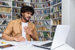 Millennial curly man in shirt sitting by desktop with portable computer and holding hand on chest. Stressed accountant experiencing heart attack symptoms and feeling hard to breath at workspace. photo