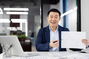 Enthusiastic Asian businessman discussing growth with a financial chart in a modern office setting. He demonstrates success and professionalism. photo