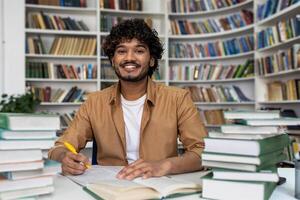 Happy and smiling student studying inside academic library, man smiling and looking at camera doing homework among books. photo