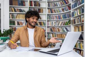 A young, curly-haired man smiles while gesturing at a laptop screen, working in a modern library setting. photo
