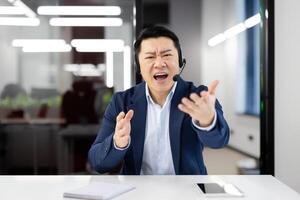 View from laptop camera of handsome asian male shouting in microphone of wireless headset while having online call through modern gadgets. Concept of technology and business. photo