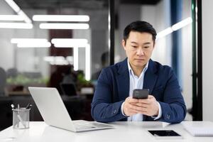 Serious Asian businessman sitting in the office at the desk with a laptop and using a mobile phone, texting, typing messages. photo