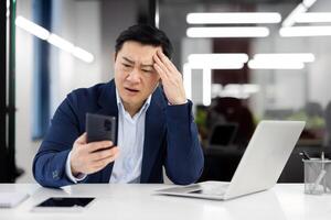 Sad disappointed asian man at workplace, dissatisfied employee in business suit with phone in hands, businessman received online notification message with bad news on smartphone, boss confused. photo