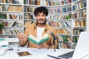 Confident blogger in white headset recording audio content while sitting with daybook by desk with professional micro. Islamic lecturer reading information for remote listeners in cyberspace. photo