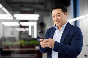 Portrait of asian adult man in blue business suit looking at smartphone screen with toothy smile on blurred background. Satisfied head of company reading pleasant text message while standing indoors. photo