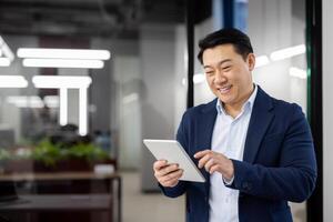 Delighted chinese head of department wearing formal suit and using digital tablet with wide smile. Happy brunette man enjoying modern technology and new opportunities for remote work. Copy space. photo
