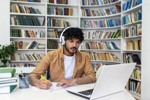 Serious concentrated man in headphones studying in library, hispanic student thinking and recording course while sitting in university campus library. photo