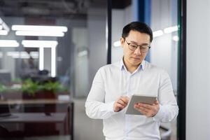 Serious thinking man with tablet computer in hands, asian man at workplace inside office, businessman in shirt reading and analyzing online data using app. photo
