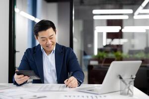A professional Asian businessman reviews financial documents while smiling in a brightly lit modern office environment, portraying efficiency and contentment. photo