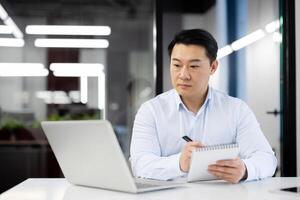 Asian business man watching training course online, man inside office at workplace writing in notebook, using laptop for remote communication with mentor and coach. photo