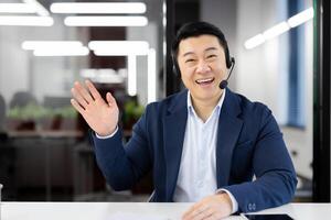 Portrait of happy asian male in business suit and headphones waving hello with hand while looking at camera. Excited man saying hi or goodbye during remote conversation with international partners. photo