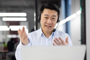 Friendly support manager in wireless headphones looking at computer screen and gesturing wit hands. Japanese distant worker taking part in meeting and making report about latest achievements. photo