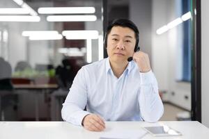 Serious brunette male in headset touching microphone with hand while looking at camera in office building. Korean hotline operator setting audio technology while sitting by desktop with tablet. photo