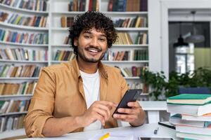 Cheerful young adult with curly hair smiling at the camera while using a smartphone, sitting at a desk with books in a library. photo