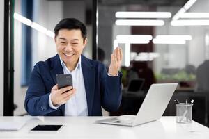 contento asiático ejecutivo atractivo en un virtual saludo, ondulación a el cámara mientras teniendo un llamada en su móvil teléfono. foto